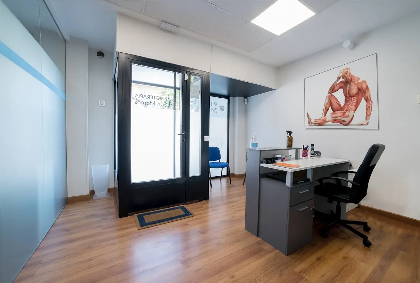 Recepción de Fisioterapia San Marcos en San Martín de la Vega, con mostrador, puerta de cristal a la calle y un póster de anatomía